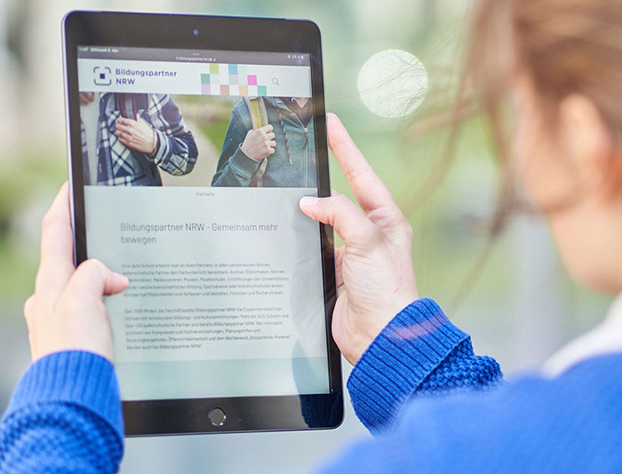 Eine Frau hält ein Tablet in der Hand. Auf dem Bidlschirm sieht man die Startseite von Bildungspartner NRW