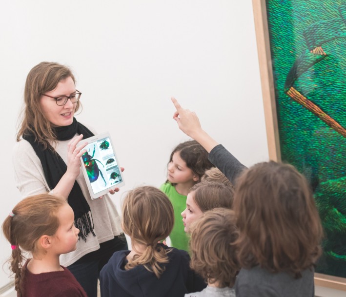 Gruppe von Kindern steht vor einem großen, grünen Bild im Museum. Eine Frau erzählt etwas und zeigt den Kindern etwas auf einem Tablet. Einige Kinder heben die Hand, um auf eine Frage zu antworten.