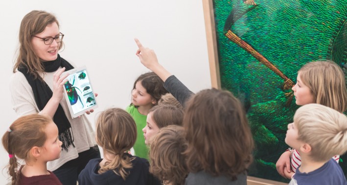 Gruppe von Kindern steht vor einem großen, grünen Bild im Museum. Eine Frau erzählt etwas und zeigt den Kindern etwas auf einem Tablet. Einige Kinder heben die Hand, um auf eine Frage zu antworten.