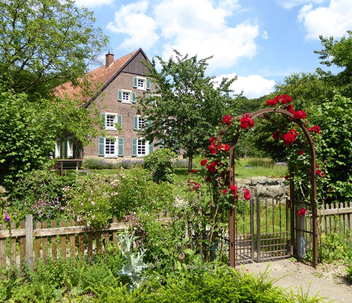 Blick vom Bauerngarten durch einen Rosenbogen auf das Haus der Biologischen Station