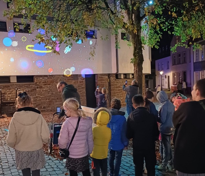 Das Foto zeigt eine Gruppe von Kindern und Jugendliche, die abends Lichtprojektionen an eine Wand werfen und diese betrachten.