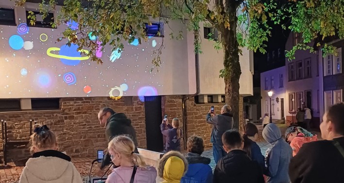 Das Foto zeigt eine Gruppe von Kindern und Jugendliche, die abends Lichtprojektionen an eine Wand werfen und diese betrachten.