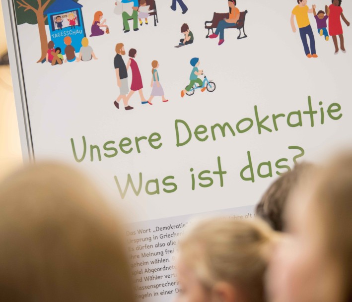 Mehrere verschwommene Hinterköpfe von Kindern vor einem Plakat mit dem Schriftzug »Unsere Demokratie. Was ist das?«