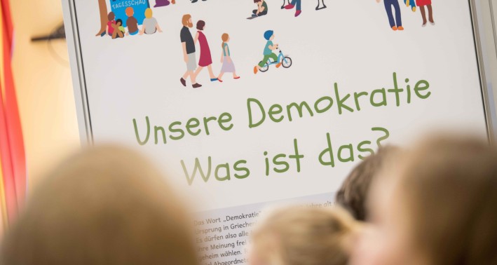 Mehrere verschwommene Hinterköpfe von Kindern vor einem Plakat mit dem Schriftzug »Unsere Demokratie. Was ist das?«