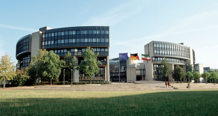 Der NRW-Landtag: ein moderner Bau geformt wie ein halvoffener Kreis. Umgeben ist das ebäude von Bäumen und Grünfläche.