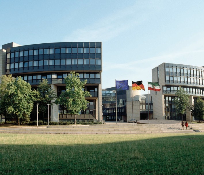 Der NRW-Landtag: ein moderner Bau geformt wie ein halvoffener Kreis. Umgeben ist das ebäude von Bäumen und Grünfläche.