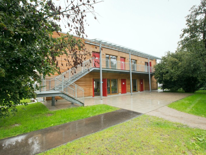 Holzgebäude mit Metalltreppe und roten Türen zwischen grünen Bäumen