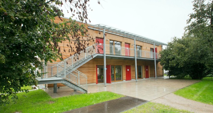 Holzgebäude mit Metalltreppe und roten Türen zwischen grünen Bäumen