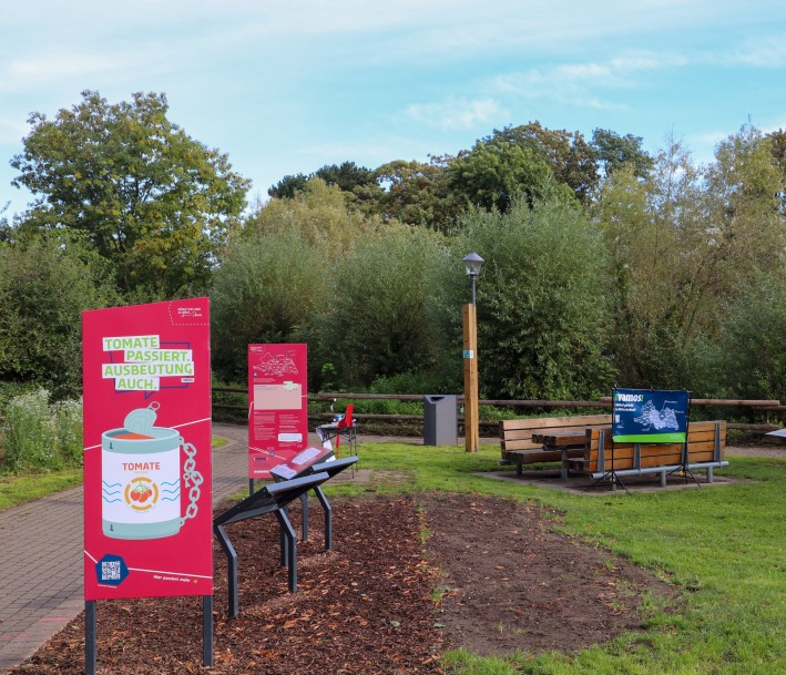 Ein Foto von einem Parkgelände mit Infotafeln und bänken. Die Infotafeln ind hauptsächlich rot gehalten und darauf steht: »Tomate passiert, Ausbeutung auch.«