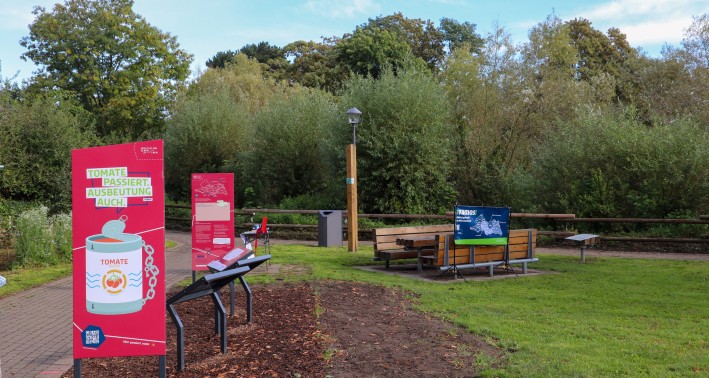 Ein Foto von einem Parkgelände mit Infotafeln und bänken. Die Infotafeln ind hauptsächlich rot gehalten und darauf steht: »Tomate passiert, Ausbeutung auch.«