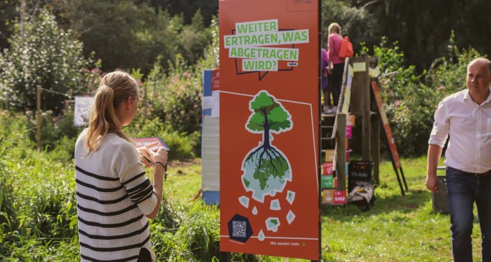 Ein Foto von einer Frau, die auf eine Infotafel schaut. Auf der Infotafel steht »Weiter ertragen, was abgetragen wird.«