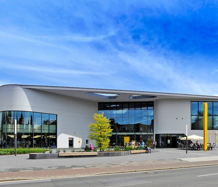 Das Bild zeigt das Hanns Dieter Hüsch Bildungszentrum. Es ist ein großes, weißes Gebäude mit einer Glasfassade und einem breiten Vordach, unter dem sich der Eingang des Gebäudes befindet.