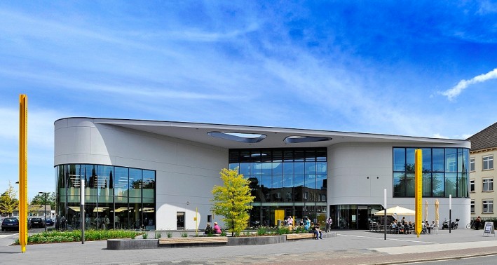 Das Bild zeigt das Hanns Dieter Hüsch Bildungszentrum. Es ist ein großes, weißes Gebäude mit einer Glasfassade und einem breiten Vordach, unter dem sich der Eingang des Gebäudes befindet.