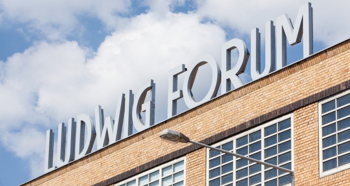 Das Bild zeigt mittig den Schriftzug „Ludwig Forum“ als Skulptur auf dem Dach des Museums. Die Gebäudeecke ist unten diagonal zu sehen, darüber ist ein blauer Himmel mit Wolken. Der Schriftzug ist weiß. Eine Straßenlaterne deutet die Höhe des Gebäudes an.
