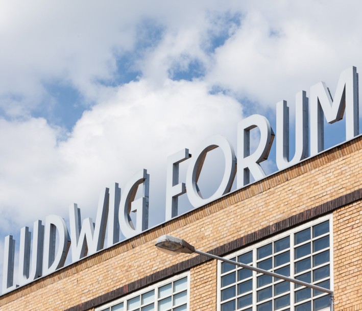 Das Bild zeigt mittig den Schriftzug „Ludwig Forum“ als Skulptur auf dem Dach des Museums. Die Gebäudeecke ist unten diagonal zu sehen, darüber ist ein blauer Himmel mit Wolken. Der Schriftzug ist weiß. Eine Straßenlaterne deutet die Höhe des Gebäudes an.
