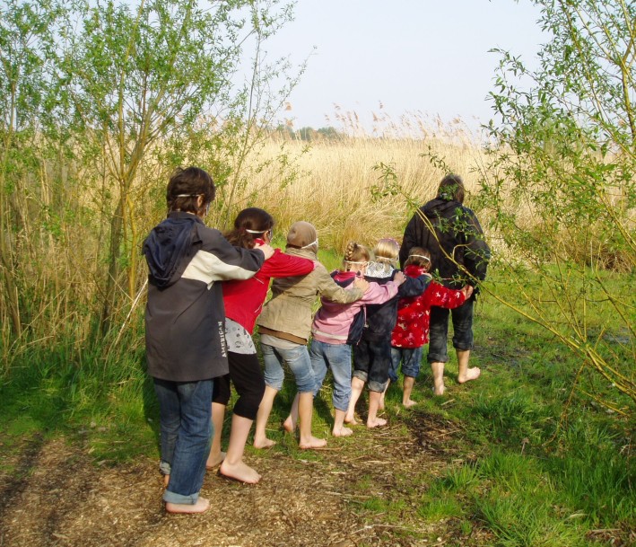 Foto: Biologische Station Rieselfelder Münster Eine Gruppe Kinder läuft aufgereiht, barfuß und mit verbundenen Augen über das Gras hintereinander her. Im Hintergrund ist eine Schilflandschaft zu sehen.