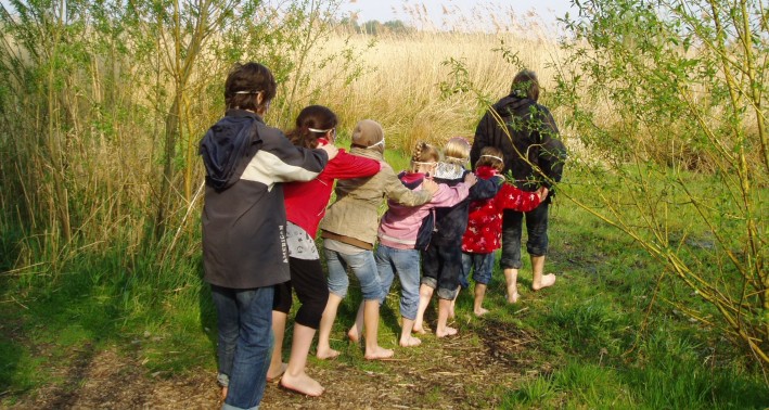 Eine Gruppe Kinder läuft aufgereiht, barfuß und mit verbundenen Augen über das Gras hintereinander her. Im Hintergrund ist eine Schilflandschaft zu sehen.