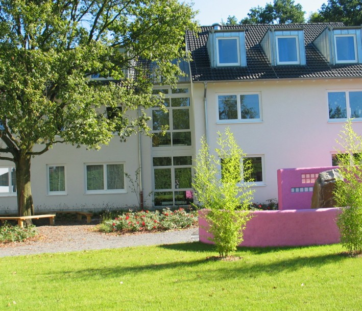 Foto: Friedensdorf International Von links nach rechts erstreckt sich im Mittel - und Hintergrund ein dreistöckiges Gebäude. Davor steht links ein Baum. Im Vordergrund ist Wiese und zwei Büsche zu sehen. s