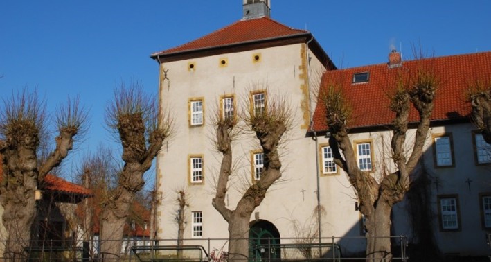 Fotografie eines Gebäudes mit heller Außenfassade und rötlichen Dach. Auf dem Dach befindet sich ein Dachreiter.