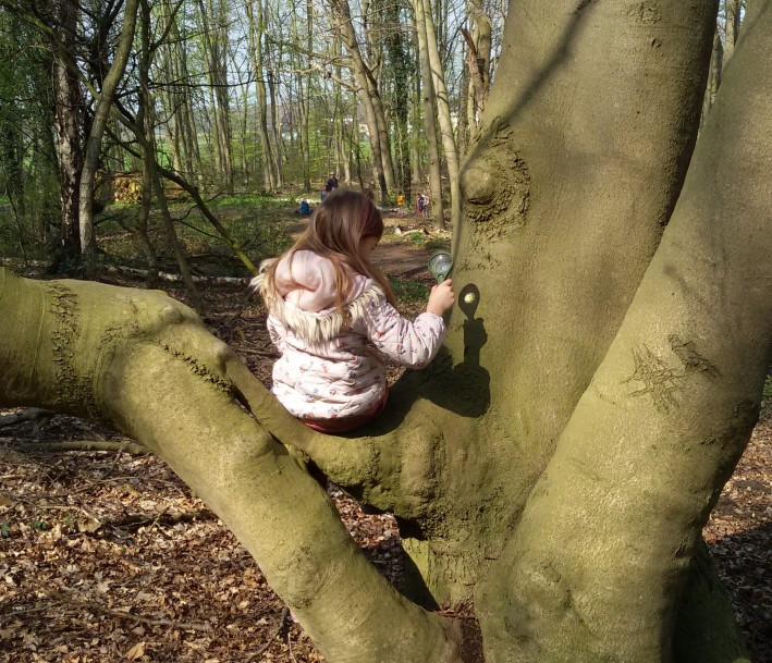 Foto: Karin Leisten Mädchen sitzt auf einem Baum trägt eine rosane Jacke und hält eine Lupe in ihrer rechten Hand