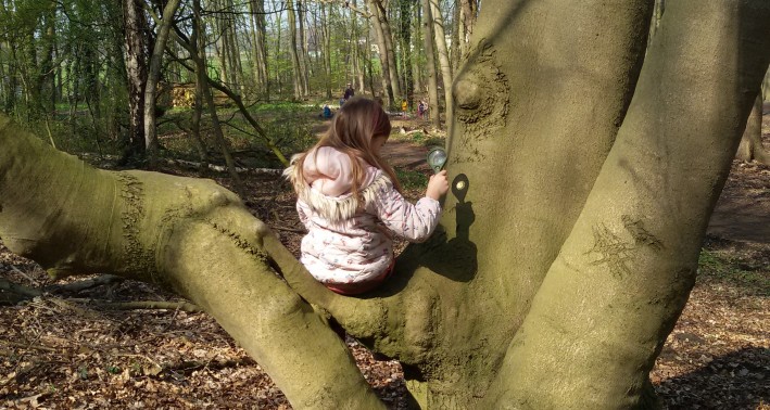 Mädchen sitzt auf einem Baum trägt eine rosane Jacke und hält eine Lupe in ihrer rechten Hand