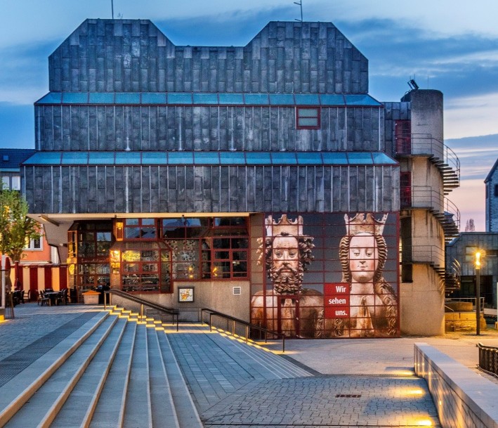 Das Bild zeigt Ein moderner Bau auf einem beleuchgteten Platz. An der Fassade ist ein Bild von zwei Reliquienbüsten des Kaiserpaares Heinrich II. und Kunigunde angebracht.
