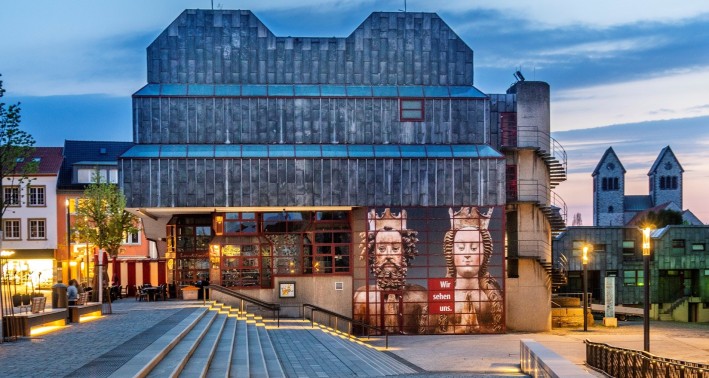Das Bild zeigt Ein moderner Bau auf einem beleuchgteten Platz. An der Fassade ist ein Bild von zwei Reliquienbüsten des Kaiserpaares Heinrich II. und Kunigunde angebracht.
