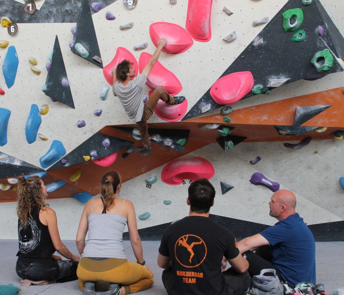 Ein Boulderer hängt an einer Boulderwand in der Boulderbar Bochum während vier Personen sitzend zuschauen