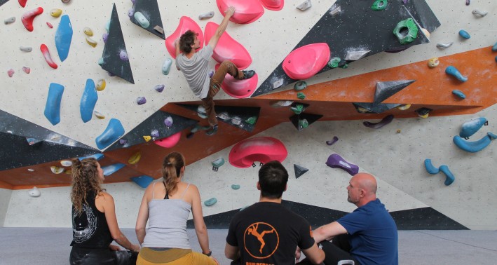 Ein Boulderer hängt an einer Boulderwand in der Boulderbar Bochum während vier Personen sitzend zuschauen