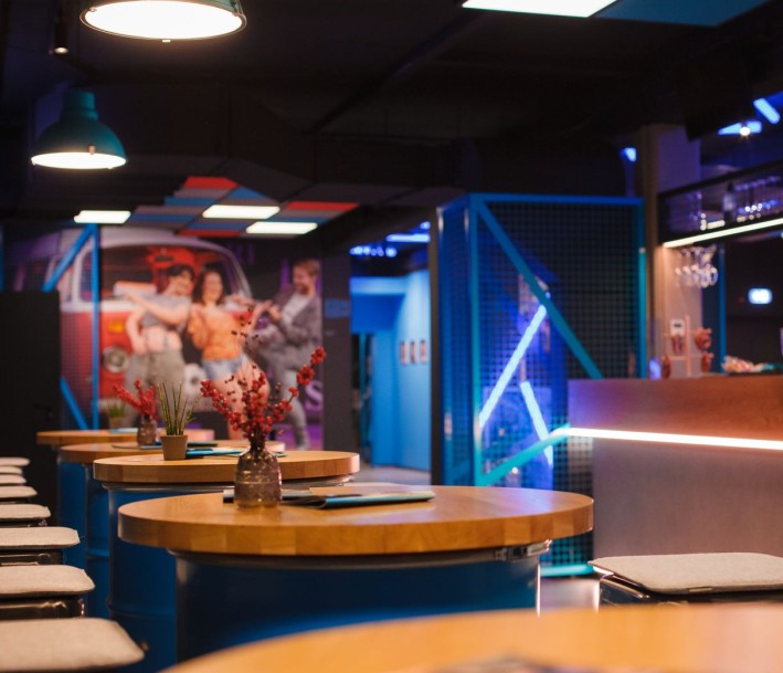 Das Bild zeigt das Forum des DAS DA THEATERS: Im Vordergrund und nach links weggehend runde Stehtische mit Hockern, rechts die große Theke, im Hintergrund an der Wand ein großes Bild der Inszenierung 