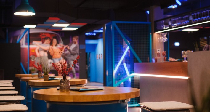 Das Bild zeigt das Forum des DAS DA THEATERS: Im Vordergrund und nach links weggehend runde Stehtische mit Hockern, rechts die große Theke, im Hintergrund an der Wand ein großes Bild der Inszenierung 'Auf und davon'.