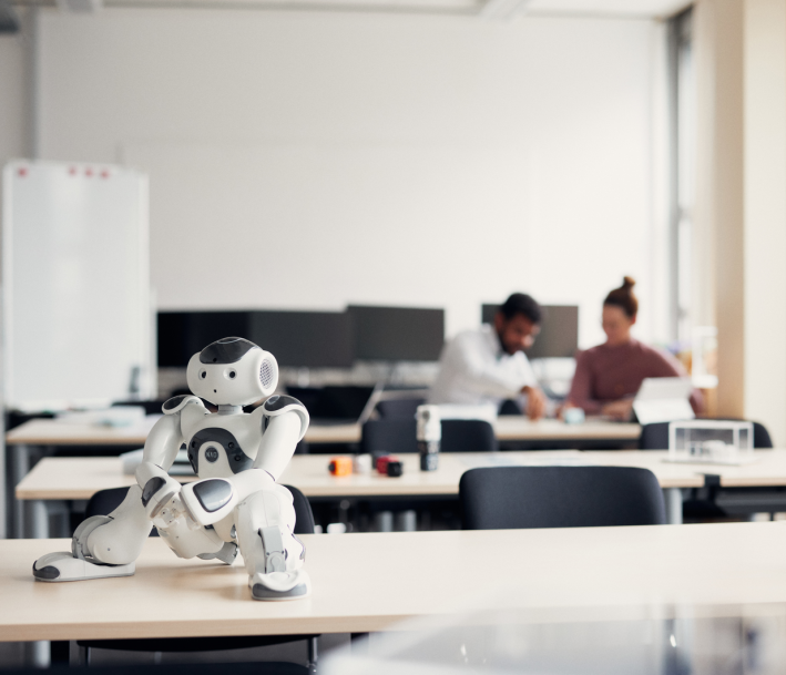 Ein kleiner weißer humanoider Roboter im Vordergrund. Im Hintergrund arbeiten zwei Menschen gemeinsam an einem Tisch im teutolab robotik der Universität Bielefeld.
