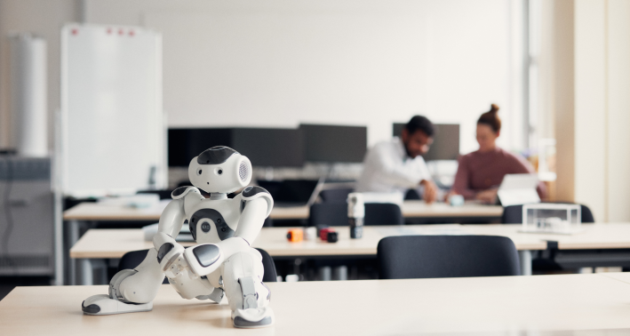 Ein kleiner weißer humanoider Roboter im Vordergrund. Im Hintergrund arbeiten zwei Menschen gemeinsam an einem Tisch im teutolab robotik der Universität Bielefeld.